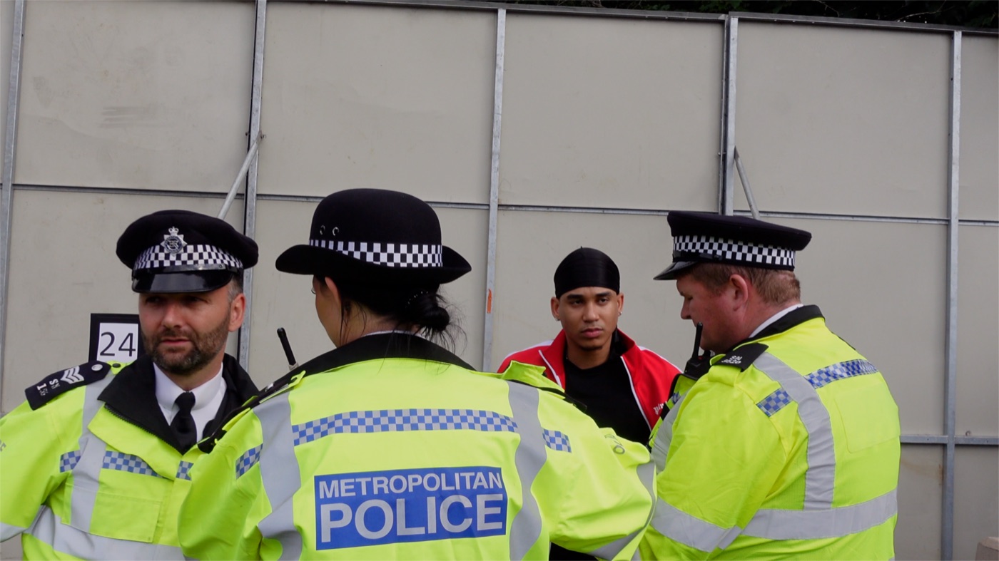 Joshua Arthur speaking with Metropolitan Police officers in daylight beside site hoarding, from the documentary Am I Really Free?