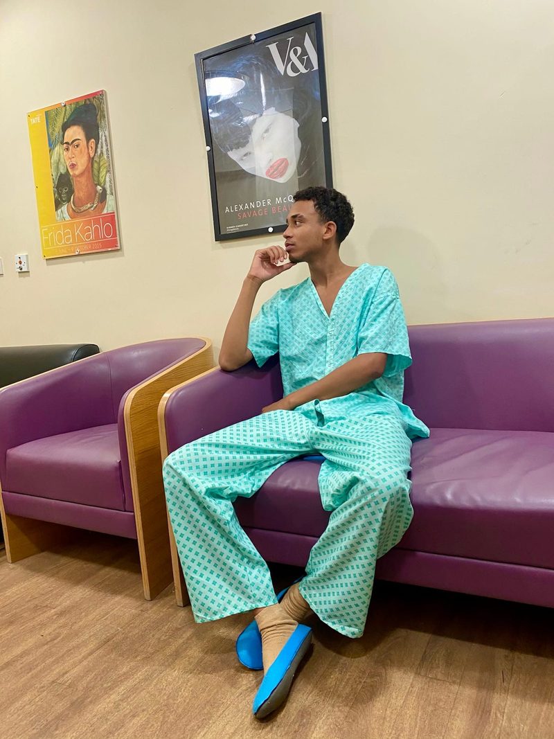 Joshua Arthur JJDREAMSS 23 years old seated in hospital waiting room Newham Centre for Mental Health London admitted 8 July 2022 wearing NHS hospital gown thoughtful Frida Kahlo Alexander McQueen Savage Beauty posters